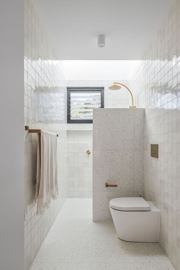 Bright, narrow contemporary bathroom with glossy white square wall tiles, a pale terrazzo floor and matching terrazzo shower partition. A wall-hung toilet sits beside the partition, with brushed brass/gold shower hardware and tap accents. The room includes a louvred window, a simple ceiling light, and a timber towel rail with a neutral striped towel.