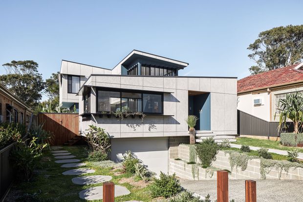 Street-facing exterior view of a contemporary multi-level residence with a strong geometric composition, pale concrete or rendered wall panels, dark-framed glazing, projecting roof planes, and a recessed covered entry. The home sits on a landscaped, gently sloping site with terraced masonry garden walls, stepping-stone paths, timber fencing, and established neighbouring houses visible on either side.