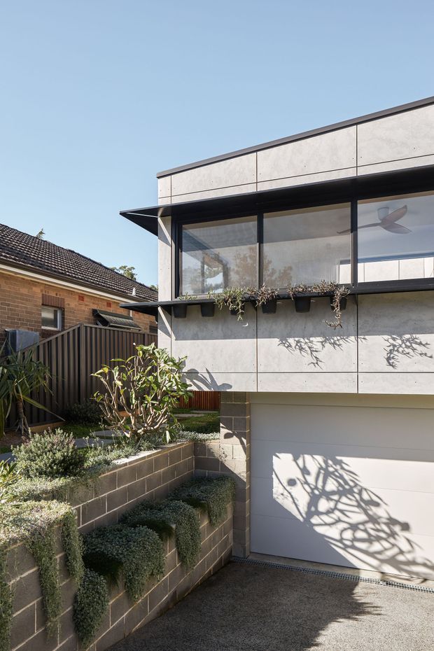 Exterior view of a contemporary two-storey residential addition beside an older brick house. The addition has a flat roof, large black-framed glazed opening, pale concrete or fibre-cement panel cladding, a projecting dark metal awning, planter boxes, and a white sectional garage door. A landscaped, terraced garden with low block retaining walls occupies the foreground, with a driveway leading to the garage.
