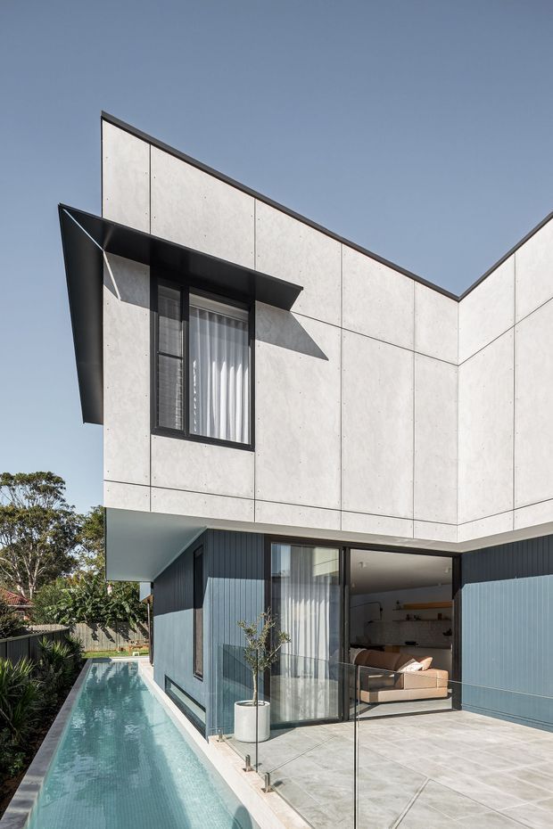 Contemporary two-storey house exterior with angular rooflines, pale concrete panel cladding, dark vertical lower-level cladding, black-framed glazing, and a narrow swimming pool running alongside a tiled terrace. The image also reveals part of an open-plan living area through the ground-floor glazing, with a sofa and built-in shelving visible.