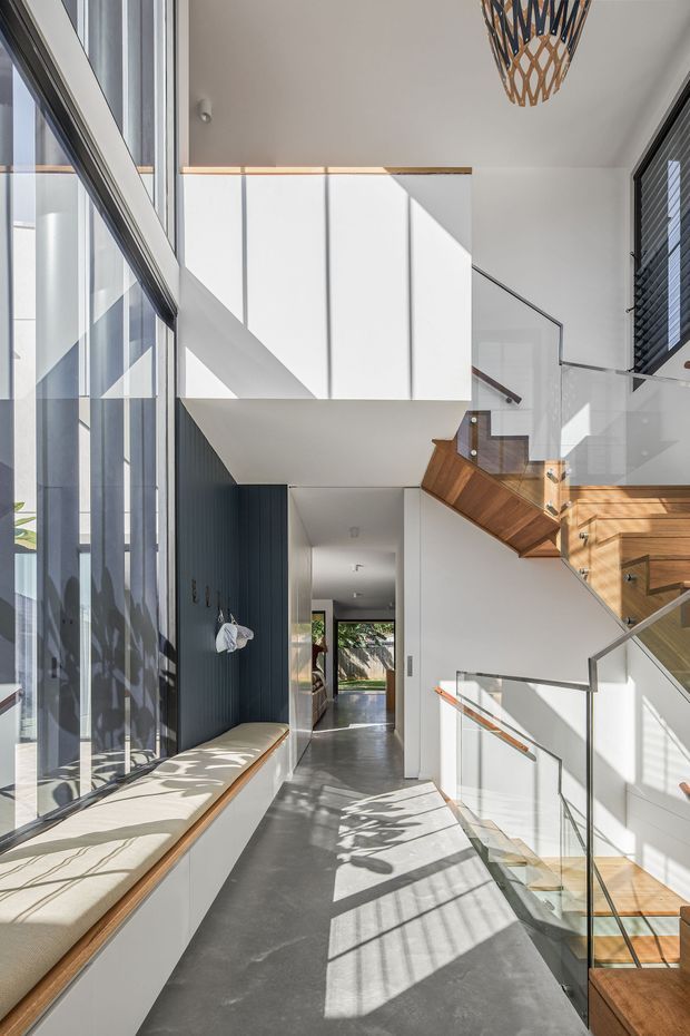Bright contemporary residential hallway and stair landing with a double-height void. Floor-to-ceiling glazing runs along the left side beside a built-in window seat, while a sculptural timber staircase with clear glass balustrades rises on the right. White walls and ceiling, dark vertical wall panelling, polished grey flooring, and strong geometric sunlight create a crisp, airy interior.