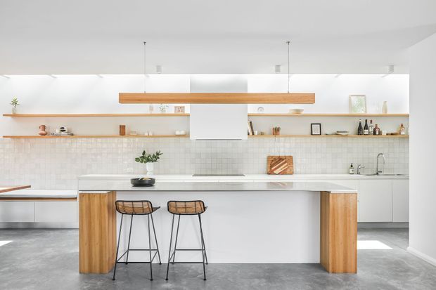 Bright contemporary kitchen with a large white island, integrated seating for two, timber end panels, open timber shelving, pale square-tile splashback, and a long timber pendant feature. The space has white cabinetry, polished grey concrete flooring, minimalist styling, and abundant natural light.