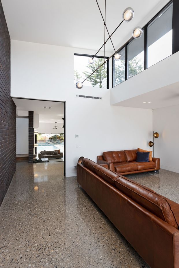 Bright double-height living room with exposed dark brickwork, polished terrazzo-style concrete flooring, white walls, and a dramatic linear pendant with globe lights. Large upper-level black-framed windows bring in abundant daylight, while brown leather sofas and sculptural floor lamps create a warm contrast. A connected dining or outdoor-facing area is visible through the doorway.