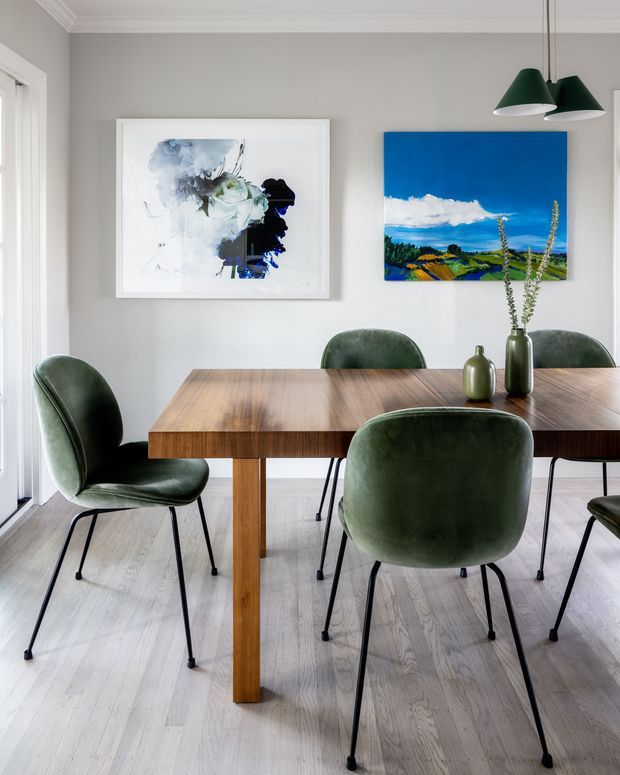 Bright, refined dining room with a rectangular timber dining table surrounded by deep green upholstered chairs on slim black legs. Light timber flooring, pale grey walls, white trim, contemporary pendant lights, and two framed artworks create a clean modern interior.