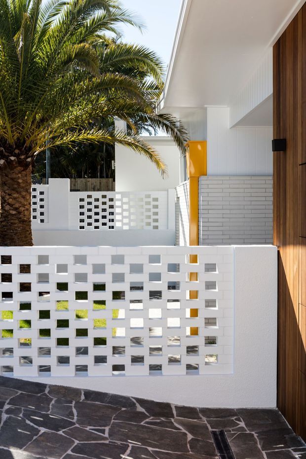 A bright, modern courtyard or entry area featuring white breeze-block privacy screens, a tropical palm, white painted brickwork and cladding, a vivid yellow accent panel, and a tall timber entrance door. The ground is finished with irregular dark stone paving, creating a strong contrast with the white architectural elements.