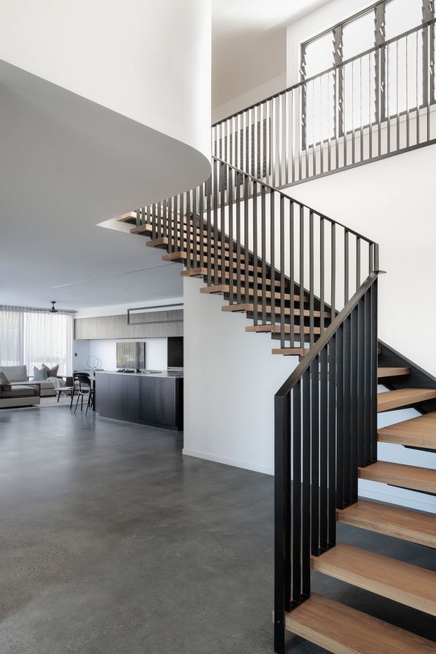 Bright contemporary open-plan interior dominated by a sculptural multi-flight staircase with timber treads and slim black metal balustrades. The stair rises beside a curved white ceiling and connects to an upper-level landing. A minimalist kitchen and living/dining area are visible in the background, with polished grey concrete flooring, white walls, dark cabinetry and large sheer-covered windows.