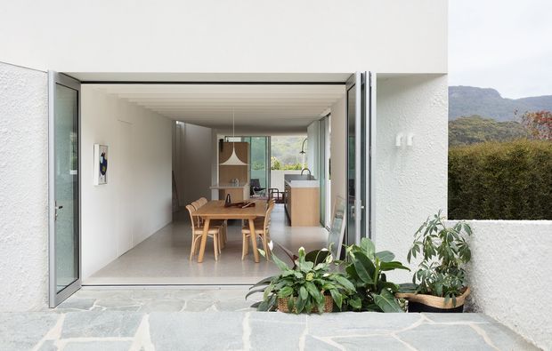 View from a pale stone-paved outdoor terrace through wide-open glazed doors into a bright, narrow open-plan interior. The space includes a timber dining table with chairs in the foreground, a compact kitchen with light cabinetry and island beyond, white textured walls, and a strongly ribbed or slatted ceiling. Potted greenery frames the opening, with a wooded hillside visible outside.