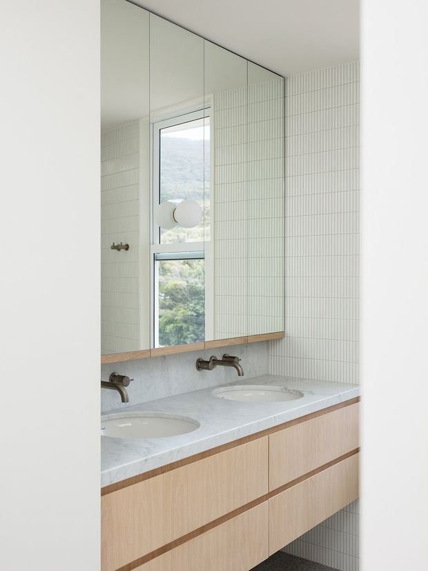 Bright contemporary bathroom vanity with a double-basin floating timber cabinet, pale stone benchtop, wall-mounted aged metal taps, and a large mirrored wall. Fine vertically lined pale tiles cover the walls, while a tall window provides views of a green hillside. The room has a clean, restrained, light-toned aesthetic.
