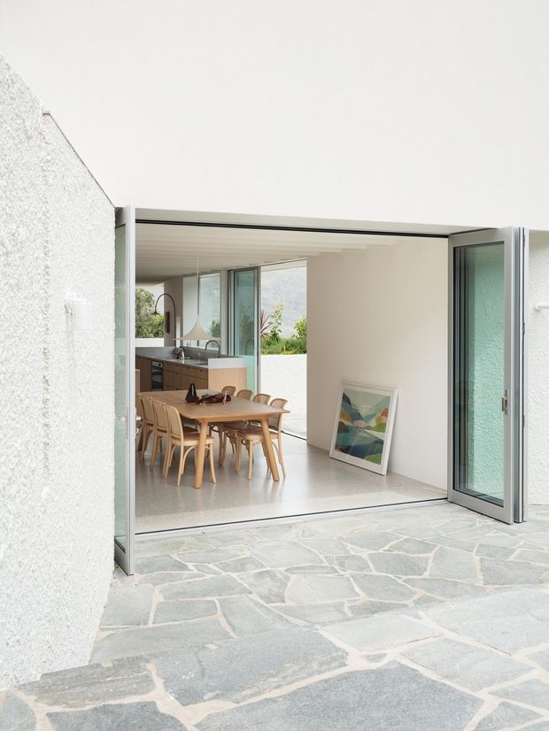 Bright contemporary courtyard view through wide-open glazed doors into an open-plan dining and kitchen area. The interior features a timber dining table with bentwood chairs, pale cabinetry, large windows, and a light polished floor. The exterior courtyard is paved with irregular grey stone slabs, while white textured walls and slim dark-framed glazing create a clean indoor-outdoor connection.