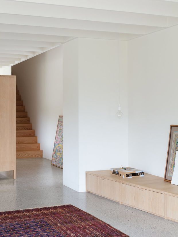 A bright minimalist interior showing a white-painted open-plan hallway and stairway. Exposed white ceiling beams run across the space, with a warm timber staircase and matching low built-in timber cabinetry along the right wall. The floor is polished grey concrete or terrazzo, complemented by a richly patterned rug, framed artwork, and a simple suspended pendant light.