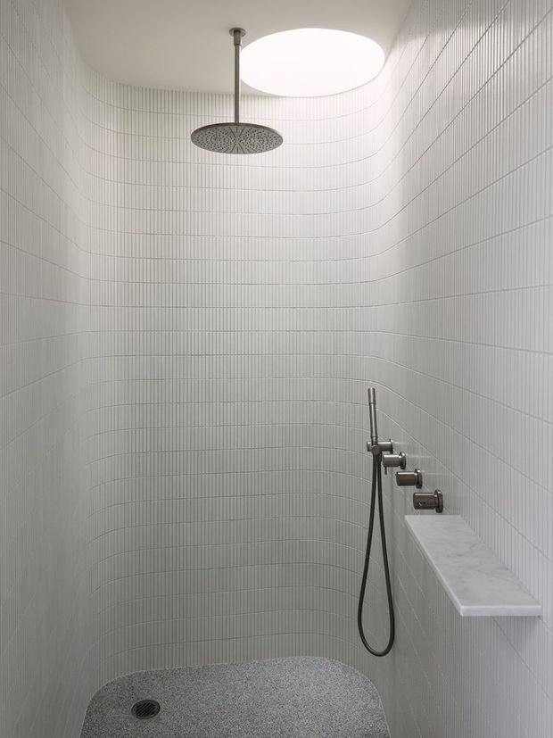 Minimalist walk-in shower enclosure with curved walls clad in fine white vertical tiles, a large round overhead rain shower, handheld shower controls, recessed marble ledge, pebble-textured floor, and a circular skylight providing soft natural light.