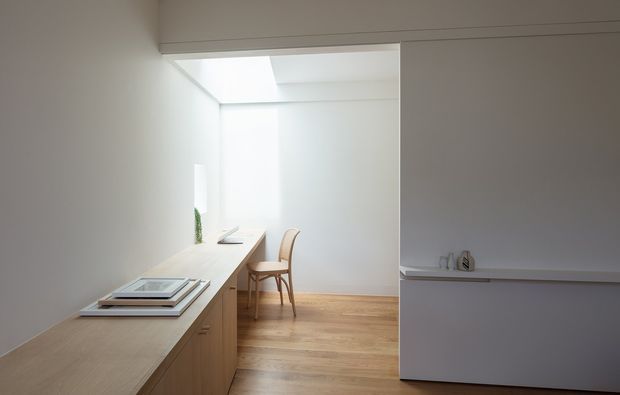 Minimalist bright interior featuring a light timber built-in cabinet and countertop extending into a compact workspace, with a simple wooden chair, white walls, pale timber flooring, and a concealed skylight or clerestory bringing in natural light. A partial-height partition with a narrow ledge separates the foreground from the workspace.