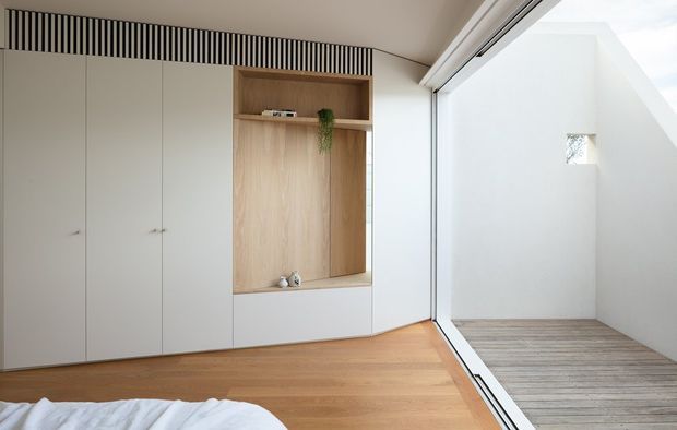Bright minimalist bedroom with floor-to-ceiling white built-in cabinetry, an integrated natural timber display niche, and warm timber flooring. A large glazed opening connects to a small enclosed timber-decked outdoor area, while a linear slatted ventilation detail adds architectural interest above the joinery.