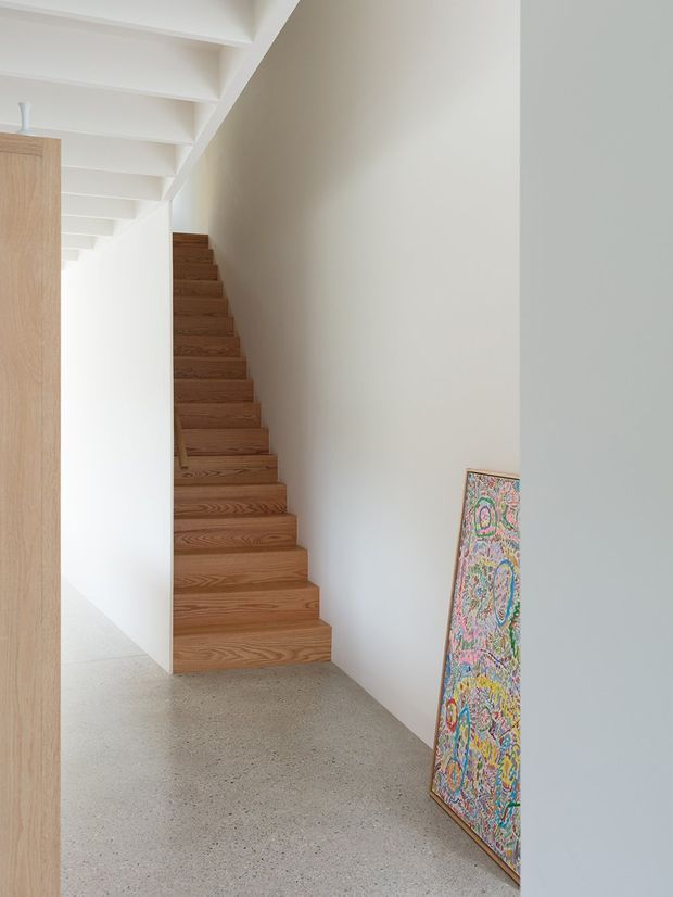 Minimalist interior hallway with a straight timber staircase tucked beneath exposed white ceiling beams. The space has white plaster walls, a light speckled concrete or terrazzo-like floor, pale timber joinery at left, and a colourful framed artwork leaning against the wall.