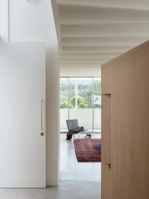 A minimalist interior passage opens toward a bright living area with full-height glazing and a leafy view beyond. The space features a white-painted exposed-beam ceiling, pale smooth walls, polished light-grey concrete flooring, and a large timber-clad built-in partition or door. A dark lounge chair and colourful woven rug add restrained accents.