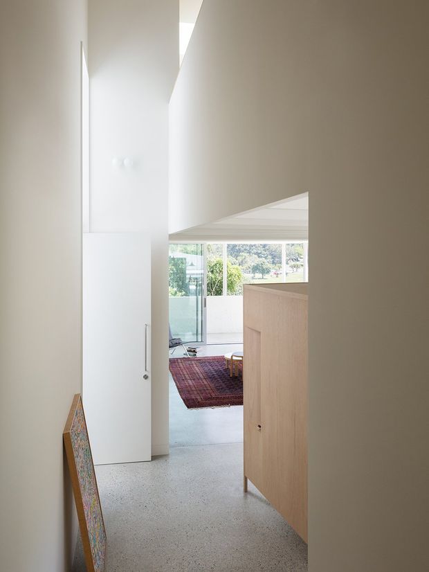 A minimalist interior view through a narrow hallway or entry passage toward a bright living area. The space features crisp white walls, a tall flush white door, a pale timber built-in cabinet or screen, polished light terrazzo flooring, and a large glazed opening overlooking greenery. A colourful rug and artwork add restrained visual accents.