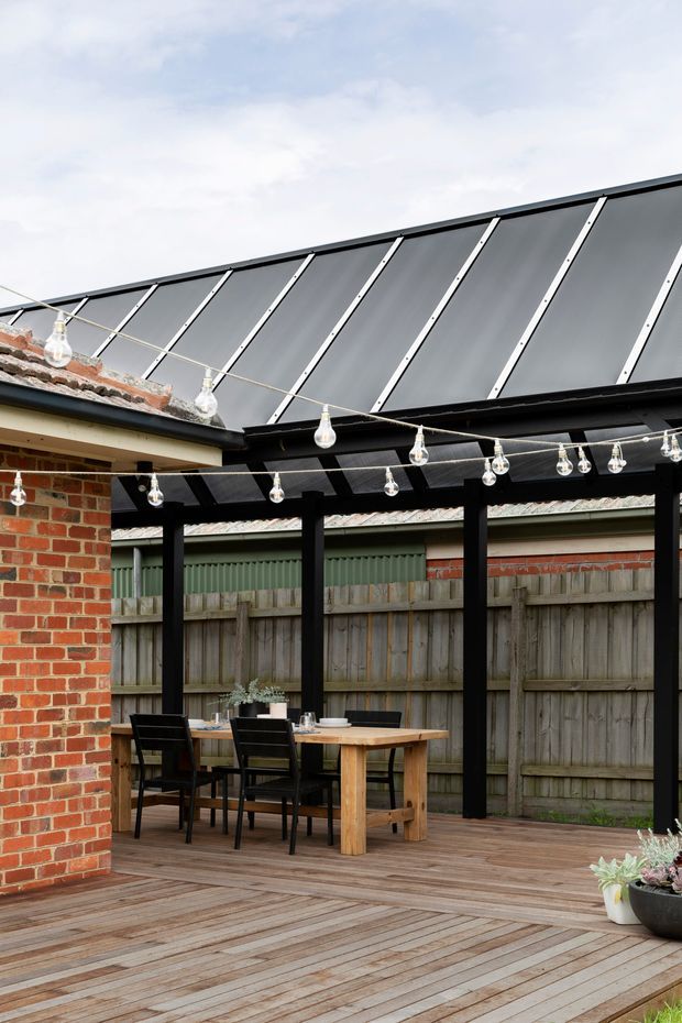A covered outdoor courtyard dining area with a timber deck, timber dining table and black chairs, framed by black-painted timber posts and beams. A sloping translucent or dark polycarbonate roof structure is visible overhead, with string lights, exposed red brick walls, and a weathered timber boundary fence. Potted plants sit at the deck edge.