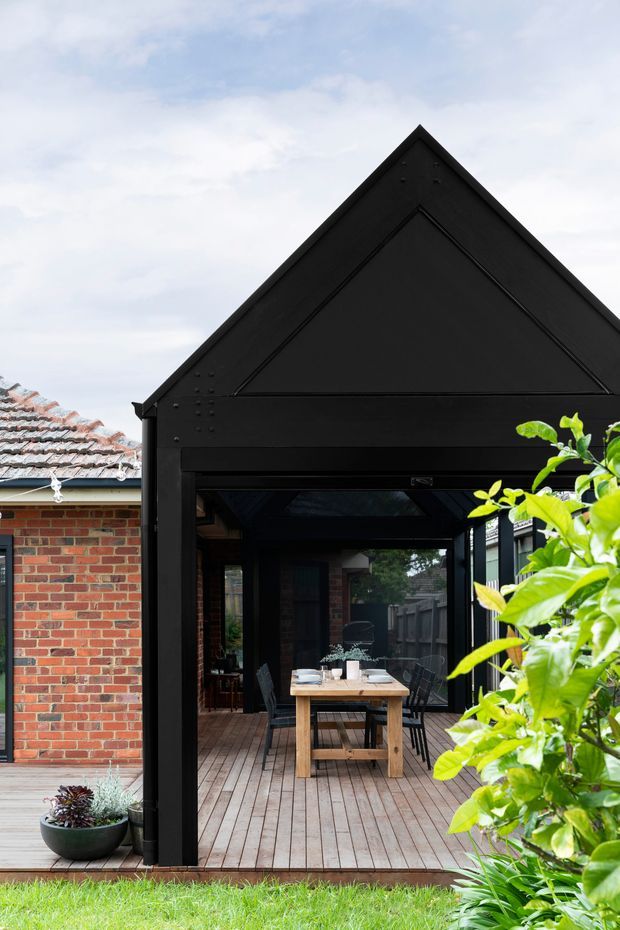Exterior view of a black-painted gable-roof extension forming a covered outdoor dining area. The enclosed timber deck has large glazed openings, a light timber dining table and chairs, and connects to an existing red-brick house with a tiled roof. Garden planting frames the foreground.