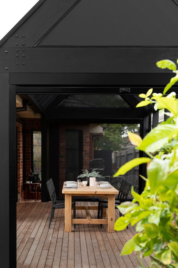 A covered outdoor dining area on a timber deck, framed by a black-painted structure with exposed riveted metal detailing and large glazed openings. A simple timber dining table is set with crockery and surrounded by dark chairs. Brick walls and a black-framed roof glazing panel are visible, with leafy planting in the foreground.