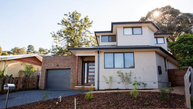 Exterior street-facing view of a contemporary multi-level suburban home with a combination of horizontal weatherboard cladding, rendered walls, and exposed brickwork. The house has a projecting upper storey, broad eaves, large windows, an attached single-car garage, a gravel driveway, and a landscaped front garden.