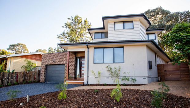Street-facing exterior view of a contemporary multi-level home with a dark low-pitched roof, cream rendered walls, horizontal weatherboard upper-storey cladding, exposed red-brick sections, large glazed windows, and an integrated single garage. The front garden has fresh mulch and young plantings, with timber fencing and neighbouring homes visible around it.