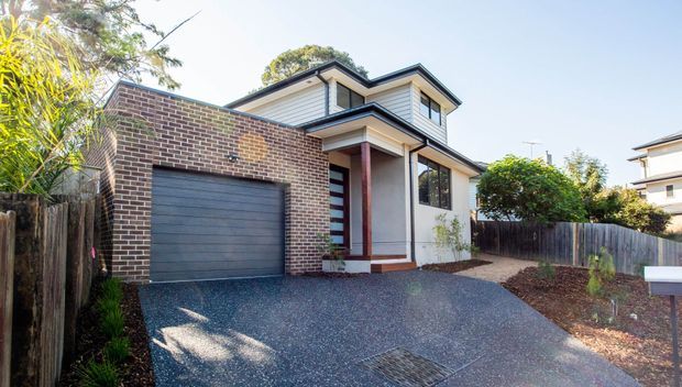Exterior view of a contemporary multi-storey townhouse with a dark brick garage wing, white horizontal cladding, projecting upper-level form, covered entry porch, dark garage door, and a paved driveway. Timber fencing and established planting frame the property.