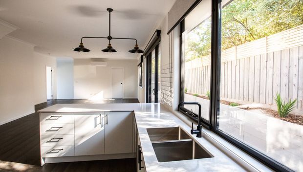 Bright contemporary kitchen with a large white island, integrated cabinetry, dark timber flooring, a double-bowl stainless-steel sink, and expansive black-framed windows overlooking a fenced outdoor area. A linear black pendant light adds contrast above the island.