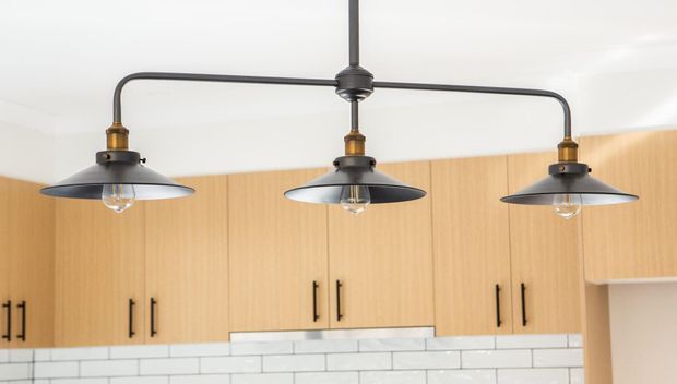Close-up interior view of a kitchen featuring warm light-wood flat-panel cabinetry with black handles, a white subway-tile splashback, and a distinctive three-light black metal pendant fixture.