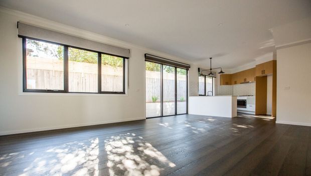 Bright open-plan living and kitchen area with dark timber-look flooring, pale walls, large black-framed windows and sliding glass doors, and a compact white kitchen island. The kitchen includes warm timber cabinetry, a white tiled splashback, and simple black pendant lighting. The room appears spacious and largely unfurnished, with natural light and views to a fenced outdoor area.