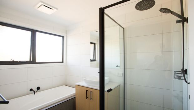 Bright contemporary bathroom with a white tiled bath surround, freestanding bathtub, timber vanity, wall mirror, and a large frameless glass walk-in shower with matte black framing and fixtures. A wide window provides natural light.