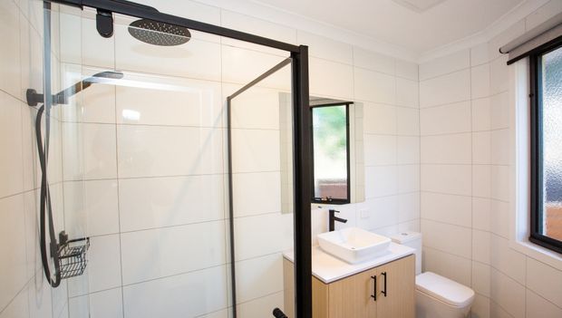 Bright contemporary bathroom featuring a frameless glass shower enclosure with black hardware, large white wall tiles, a timber-look floating vanity with white vessel basin, black tapware, toilet, and a frosted-glass window.