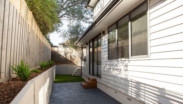 A narrow side courtyard beside a contemporary single-storey residence, featuring light horizontal weatherboard cladding, extensive dark-framed glazing and sliding doors, a timber entry stair, paved walkway, raised planter bed, vertical timber boundary fencing and a small strip of lawn.
