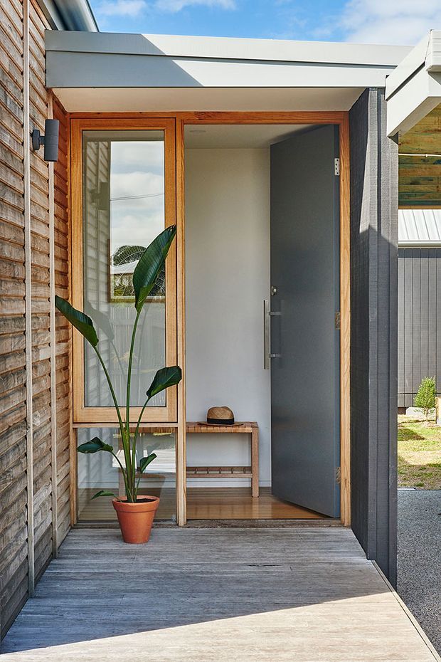 A bright contemporary residential entryway viewed from outside, with a covered porch and open dark-grey front door. The entrance features warm timber-framed glazing, horizontal timber wall cladding, grey timber decking, a simple built-in timber bench, and a potted tropical plant. Clean rectilinear detailing and strong natural light create a welcoming modern facade.
