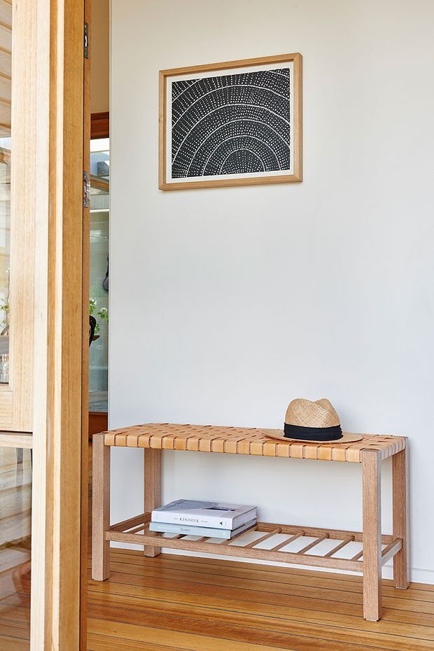 A bright, calm entryway featuring a light timber console bench with a slatted lower shelf, positioned against a pale painted wall. A framed black-and-white artwork hangs above, while a timber-framed opening and glimpses of adjacent built-in shelving are visible at left. The floor and surrounding joinery are also timber, creating a warm natural finish.