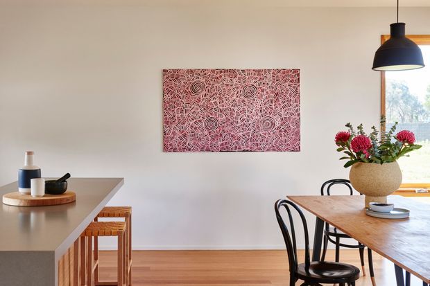 A bright, warmly styled open-plan dining and kitchen area. A timber dining table with curved black chairs sits beside a pale island or peninsula with timber stools. Light painted walls, timber flooring, a black pendant light, and a colourful abstract artwork create a refined contemporary interior, with greenery visible through the window.