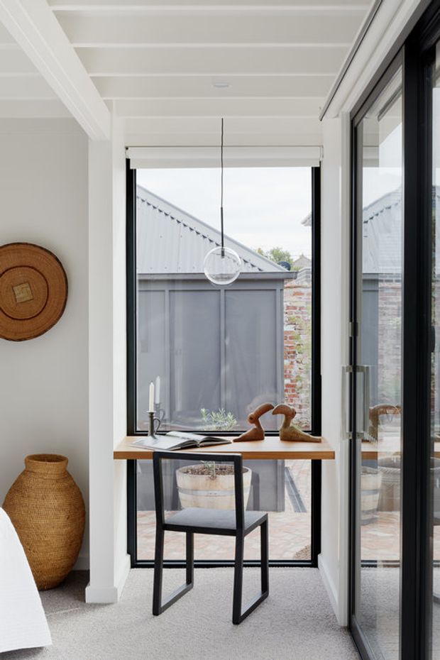 A bright, compact interior workspace or reading nook beside large black-framed glazed doors and a window. The space features a slim timber desk with a black chair, a suspended spherical pendant light, white timber-lined ceiling and walls, pale carpet, a woven floor basket, and natural decorative objects. A small paved courtyard and adjoining outbuilding are visible through the glazing.