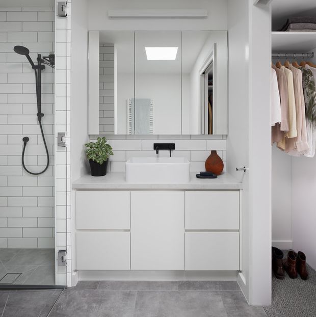Bright, compact contemporary bathroom with a walk-in tiled shower, wall-mounted white vanity, rectangular basin, mirrored cabinet, and matte black tapware. The bathroom opens directly to a walk-in wardrobe visible on the right, with hanging clothes and shoes. Grey floor tiles, white subway wall tiles, and integrated storage create a clean, efficient layout.