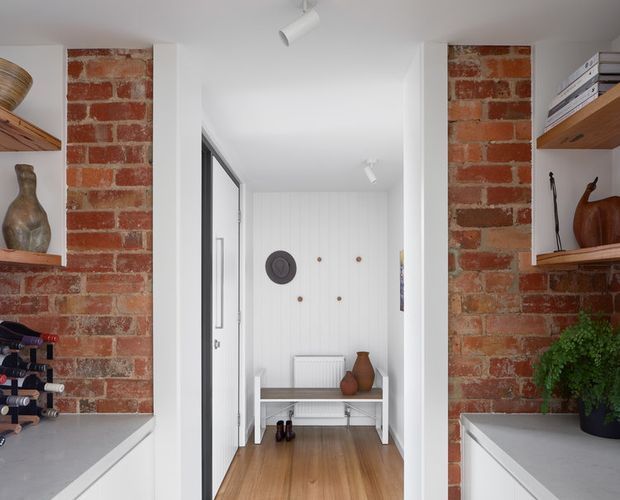 Bright contemporary entryway viewed from an adjacent space, framed by exposed red brick walls and white built-in shelving with decorative objects and a small wine rack. The narrow hallway has warm timber flooring, white vertical wall panelling, a simple bench, coat hooks, a radiator, and recessed-style track lighting. Black-framed glazing or a door is visible along the left side.