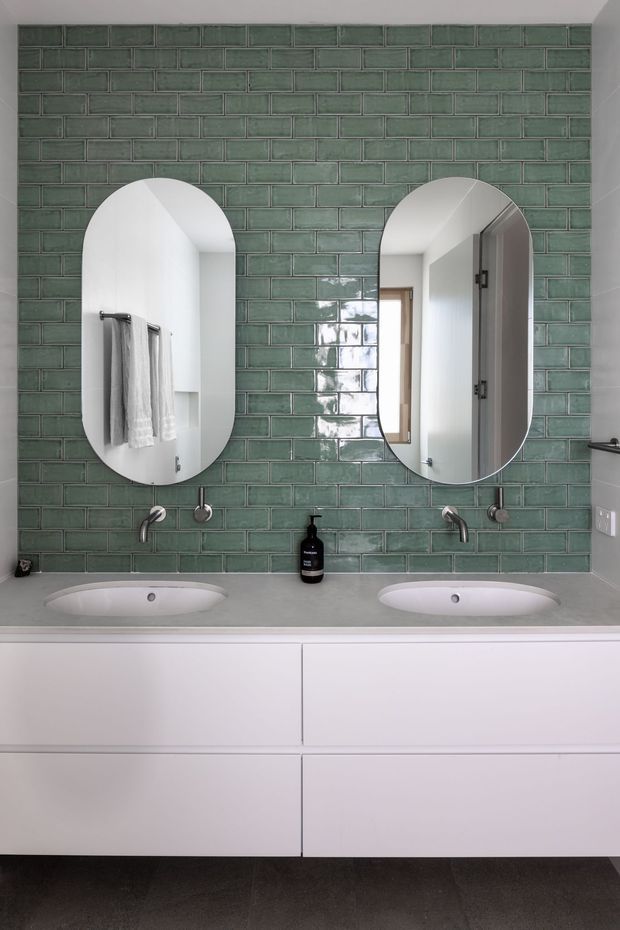 A contemporary bathroom vanity with two integrated white basins, brushed metal wall-mounted taps, and paired rounded-arch mirrors. The main feature is a full-height glossy green subway-tile backsplash with dark grout, set above smooth white handleless cabinetry and a grey benchtop. The mirrors reflect a towel rail, doorway, and light-filled adjacent space.