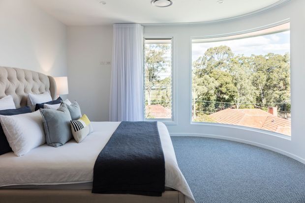 Bright, serene bedroom with a tufted upholstered bedhead, layered neutral bedding, and a dark charcoal bed runner. The room features pale walls, grey carpet, full-height curtains, recessed ceiling details, and a curved bay window overlooking leafy suburban surroundings and terracotta rooftops.