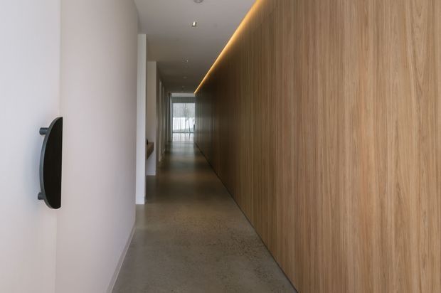 A long, narrow contemporary hallway with a smooth light wall on the left and full-height warm timber wall panelling or cabinetry on the right. A recessed linear LED strip runs along the top of the timber wall, while a polished concrete floor leads toward glazed doors or windows at the far end. Minimal black wall-mounted hardware provides a small contrast.