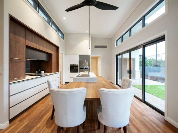 A bright contemporary open-plan kitchen and dining area with a long timber dining table, upholstered dining chairs, a white island, and extensive built-in cabinetry. The room has warm timber flooring, dark timber display and media joinery, white cabinetry, high ceilings, clerestory windows, a black ceiling fan, and large glazed sliding doors opening to a small lawn and paved outdoor area.