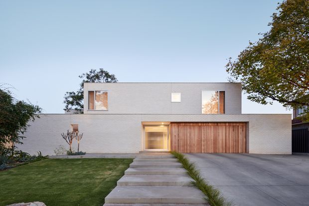 Front exterior view of a minimalist two-storey residence with a pale rendered or masonry facade, flat roofline, large rectangular upper-level windows, a warm timber garage door, and a concrete path leading from the lawn to the recessed entry. A broad driveway and landscaped front garden complete the composition.