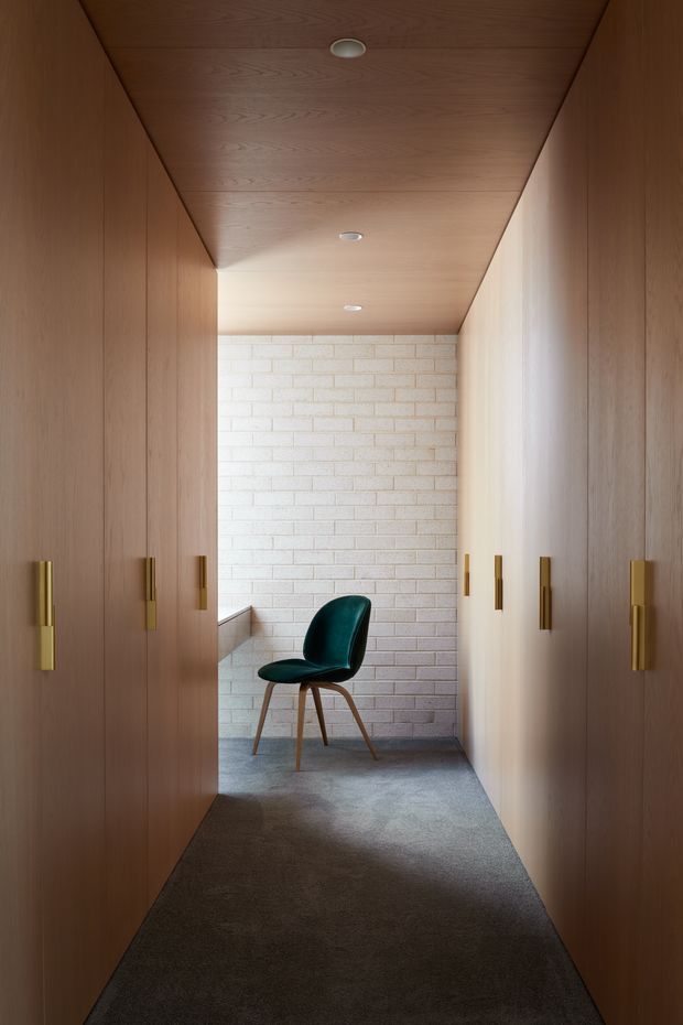 A narrow, richly timber-lined corridor or walk-in wardrobe with full-height wood cabinetry on both sides, brass vertical handles, a timber ceiling with recessed downlights, and a pale exposed brick feature wall at the end. A single dark teal upholstered chair sits on a grey carpeted floor, creating a refined, warm and restrained interior.