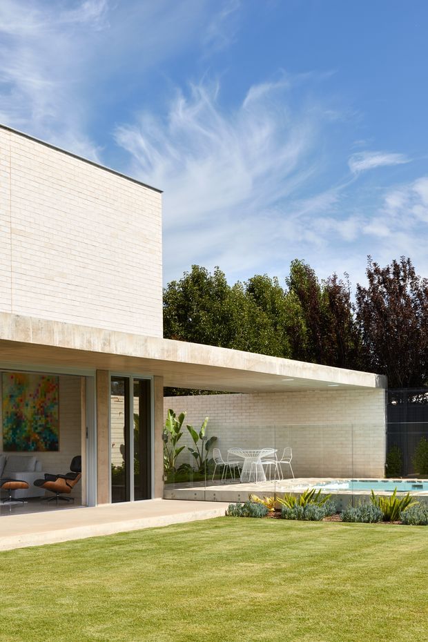 Exterior view of a modern single-storey residence with pale brick masonry walls, a broad flat concrete roof or canopy, extensive indoor-outdoor glazing, a covered patio with outdoor dining furniture, landscaped garden beds, and a swimming pool. The image also reveals part of a refined living room through the open glazing.