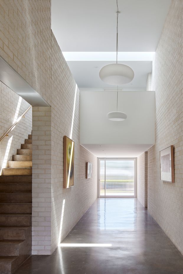 A bright, elongated interior hallway with pale exposed brick walls, a polished concrete floor, and a dark concrete stairway ascending along the left side. Large circular pendant lights hang beneath a high white ceiling with a linear skylight or clerestory opening. Framed artwork lines the walls, and a glazed door at the far end brings in daylight. The space has a minimalist, institutional-industrial character with strong geometric lines and abundant natural light.
