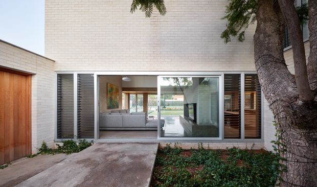 A low-rise contemporary residential facade viewed from an entry path, with pale brick walls, a timber garage or entry door, and broad aluminium-framed glazing opening onto a living area. Vertical louvres flank the glazing, while a mature tree and planted bed soften the paved approach. The interior visible through the glass includes a sofa and timber-lined spaces.