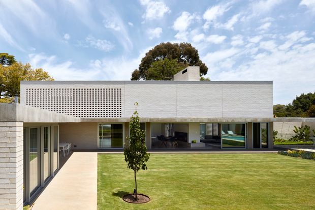 A low-slung modern residential house viewed from the landscaped front garden. The building has a long pale textured masonry facade, a flat roof, an upper perforated breeze-block screen, broad floor-to-ceiling glazing, and a sheltered outdoor living area opening onto a rectangular lawn. A pale paved path and planted borders frame the minimalist composition beneath a bright sky.