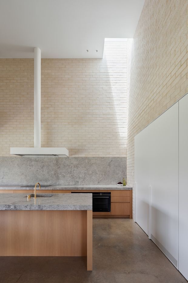 Bright contemporary kitchen with a large timber island, grey stone benchtops and splashback, integrated appliances, a brass tap, white cabinetry, pale exposed brick walls, polished concrete flooring, and a high ceiling with clerestory-style glazing or skylight. The space has a clean, refined material palette and strong natural light.