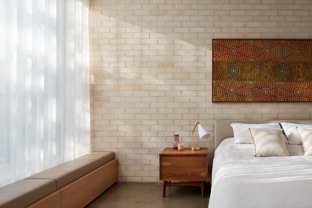 A softly lit bedroom with a warm pale brick feature wall, floor-to-ceiling sheer curtains, a built-in timber window seat, and a neatly made bed. A mid-century-style timber bedside table, angled table lamp, and framed patterned artwork add warmth and character. The room has a restrained, natural palette and a calm contemporary feel.